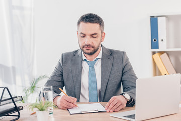 Naklejka premium handsome businessman in suit sitting at desk and writing in office