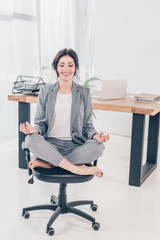 Beautiful smiling businesswoman in suit sitting on chair and meditating in Lotus Pose in office