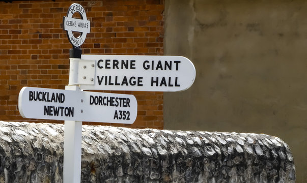 Public vintage metal direction sign, in Dorset. Post standing in front of faced flint stone wall. Brick and rendered background. Cerne Abbas. England.
