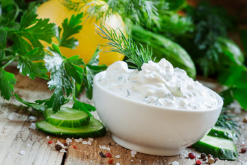 White greek sauce in white bowl with cucumber, lemon juice, herbs and spices on old wooden table, selective focus
