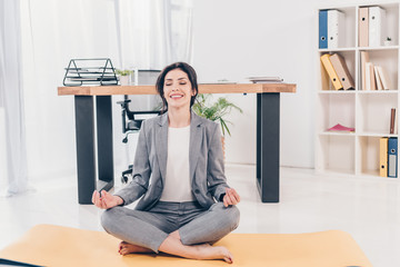 beautiful smiling businesswoman in suit sitting on fitness mat and meditating in office