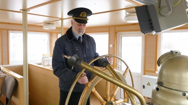 Elderly grey-haired captain of the ship in peaked cap at the helm in the wheelhouse is talking about his job. Captain at the steering wheel of the ship