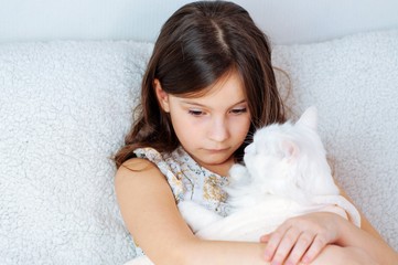 Home portrait of a little cute girl with a white cat Felis catus with blue eyes on a soft sofa