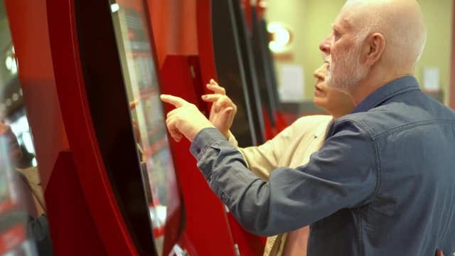 Senior couple choosing movie and buying ticket from vending machine at movie theater at mall via touch screen. Senior man making gestures by touching screen. Active modern life after retirement.