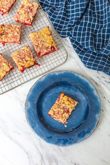Freshly Baked Strawberry Bar Cookies cooling on a wire rack, one isolated in front on blue plate, blue kitchen towel, white countertop