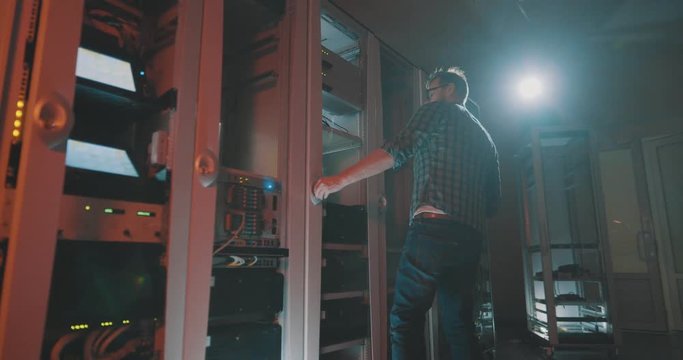 Side view of young programmer adjusts work of server in server cabinet while working with supercomputer in data center