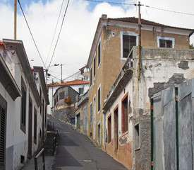 a narrow hillside street in funchal madeira with old Portuguese houses with shutters with a blue...