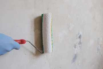 A hand in a blue glove holds a roller for finishing work. The process of priming the white wall....