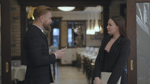 Portrait male and female business partners meet in the restaurant. People shaking hands. Lady in an elegant suit coming to the bearded man waiting for her near the entrance