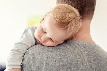 Portrait of cute adorable blond caucasian toddler boy sleeping on fathers shoulder indoors. Sweet little child feeling safety and care on daddys hand. Single father concept