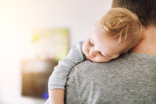 Close-up portrait of cute adorable blond caucasian toddler boy sleeping on fathers shoulder indoors. Sweet little child feeling safety daddys hand. Responsibility and childcare. happy childhood