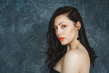 Fototapeta premium Fashion portrait of a stylish brunette girl wearing a black dress. Woman with long hair wearing earrings. Set on a gray background. A look at the camera