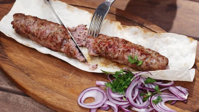 Close-up of grilled lula kebab, traditional oriental dish, woman cuts with knife and fork in restaurant