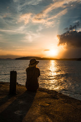 Fototapeta premium silhouette of a Girl in straw hat sitting in front of ocean and sunset cloudy blue and orange sky