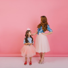 Full-length portrait of slim young mother and charming daughter wearing the same trendy outfit, posing at party. Smiling curly young woman gently holds little stylish girl in white crown by chin.