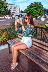 A brunette girl sits in the summer on a bench. In the hands of phone, makes photos on the smartphone. Happy smiling in sunglasses, green blouse and white skirt shorts.