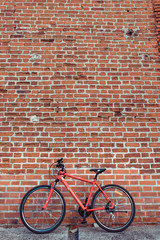 Naklejka premium Red bicycle on the background of a brick old wall. Free space for text. The concept of parking and abandoned bike. Locked and parked, locking cable on the frame of the bike. In summer in the city.