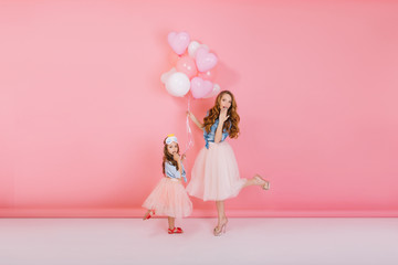 Graceful young mother funny dancing with her little daughter waiting for guests at birthday party. Adorable sisters holding balloons and posing with surprised face expression on the pink background