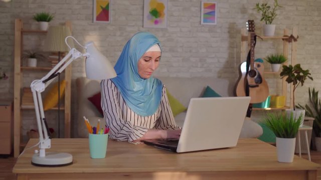 Young muslim woman in a hijab with a scar from a burn on her face works at a laptop