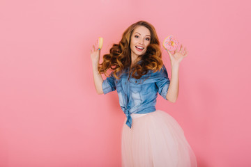 Cheerful curly girl in trendy skirt holding delicious donuts and rejoices at the end of diet. Portrait of jumping long-haired woman in retro outfit, posing with sweets isolated on pink background