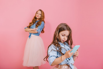 Young adorable woman  looks with a smile at her little curly daughter playing in the app on phone. Charming girl in trendy outfit gladly posing while her kid sister texting message on pink background