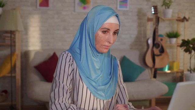 Portrait young muslim woman in a hijab with a scar from a burn on her face works at a laptop
