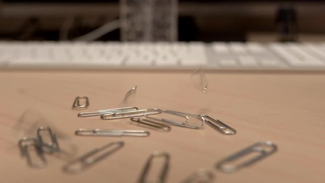 A hand full of paper clips slowly dropping onto an office desk in slow motion a little bit at a time.