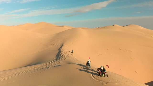 Aerial of desert sand dunes with motocross riders and a ​man running.