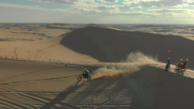 Aerial of desert sand dunes with motocross riders and a ​man running.