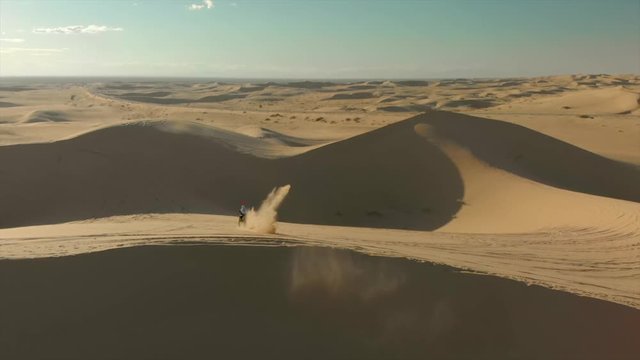 Aerial of desert sand dunes with motocross riders and a ​man running.