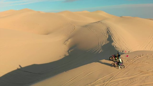 Aerial of desert sand dunes with motocross riders and a ​man running.