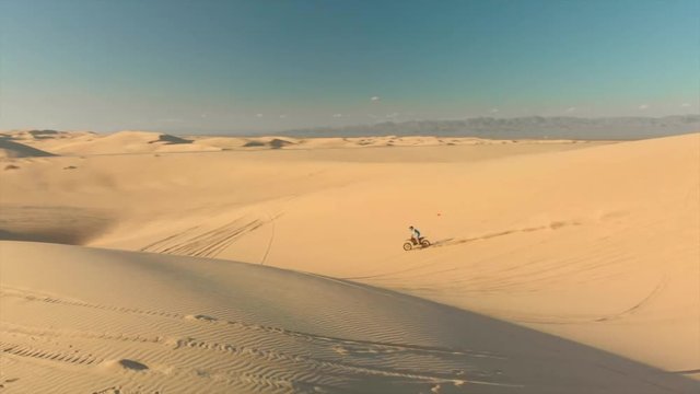 Aerial of desert sand dunes with motocross riders and a ​man running.