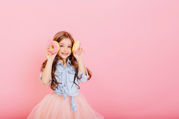 Obraz na płótnie Canvas Cute little girl with long brunette hair having fun to camera with donuts isolated on pink background. Charming fashionable princess in tulle skirt expressing positivity. Place for text