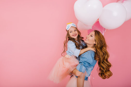 Adorable Laughing Birthday Girl With Colorful Balloons Embracing Her Young Smiling Mom After Funny Event. Attractive Mother Posing With Pretty Daughter In Mask At Party Isolated On Pink Background