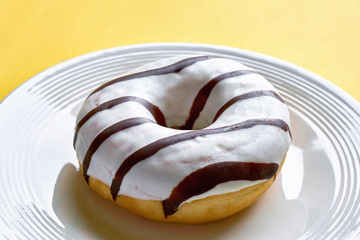 One single round donut with white ice glazing and stripes of dark brown chocolate on a white plate against a yellow background with copy space. 