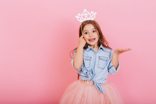 Joyful little girl with long brunette hair in tulle skirt holding princess crown on head isolated on pink background. Celebrating brightful carnival for kids, expressing positivity of birthday party