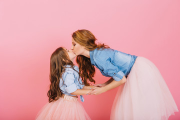 Stylish curly mom and daughter hold hands and kiss sweetly at children's event on the pink background. Little long-haired girl in denim shirt and lush skirt kissing her young mother at birthday party