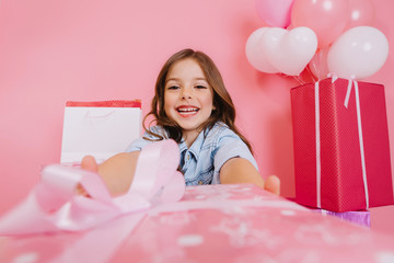 Closeup pink present giving little joyful girl to camera on pink background. Smiling suround big giftboxes, balloons, celebrating birthday party, expressing positivity