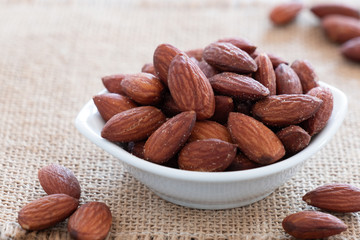 many Almonds in white bowl put on brown mat background light from back.