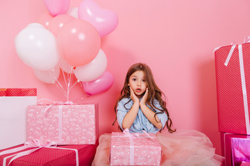 Portrait sweet surprised birthday kid looking to camera suround a lot of giftboxes, balloons isolated on pink background. Cute little girl with long brunette hair expressing with present on knees
