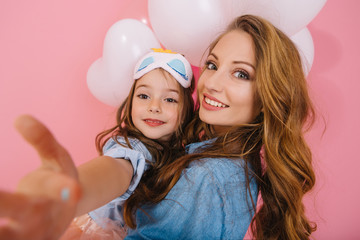 Close-up portrait of beautiful young woman in denim shirt holding her charming little daughter at birthday party. Pretty curly smiling sisters posing with white helium balloons on pink background
