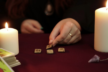 Witch is fortune teller in black mantle cast a spell on ancient runes. Tarot cards, amethyst stones, white candles on dark mystic background. Occult, esoteric, divination and wicca concept.