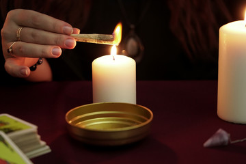 Witch is fortune teller in black mantle with rings conjures burning paper with a spell. Tarot cards, amethyst stones, white candles on dark background. Occult, esoteric, divination and wicca concept.