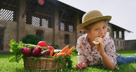 Portrait of a happy little girl is eating fresh just harvested vegetables and smiling in camera on a background of a countryside farm.