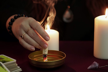 Witch is fortune teller in black mantle make ritual and burns paper with spell. Tarot cards, amethyst stone, white candles on dark mystic background. Occult, esoteric, divination and wicca concept.