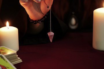 Witch is fortune teller in black robe holds in hand amethyst pendulum on chain. Tarot cards, amethyst stone, white candles on dark mystic background. Occult, esoteric, divination and wicca concept.