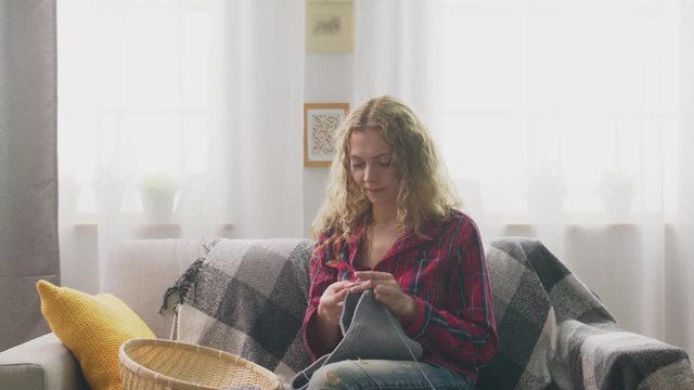 Zoom in of woman sitting on couch and knitting in home and looking in camera