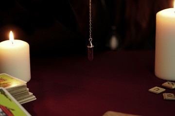 Tarot cards, amethyst magic stone pendulum on a chain, white candles and ancient wooden runes on dark mystic and vintage background. Occult, esoteric, divination and wicca concept.