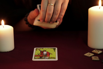 A witch is fortune teller in a black mantle pulls a tarot card from deck. Tarot cards, ancient runes and white candles. Occult, esoteric, divination and wicca concept. Mystic and vintage background.