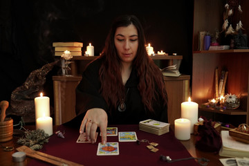 Witch is fortune teller in robe performs a rite of black magic. Magical ritual. Tarot cards, white candles, aroma stick, ancient book and runes. Occult, esoteric, divination concept. Mystic background